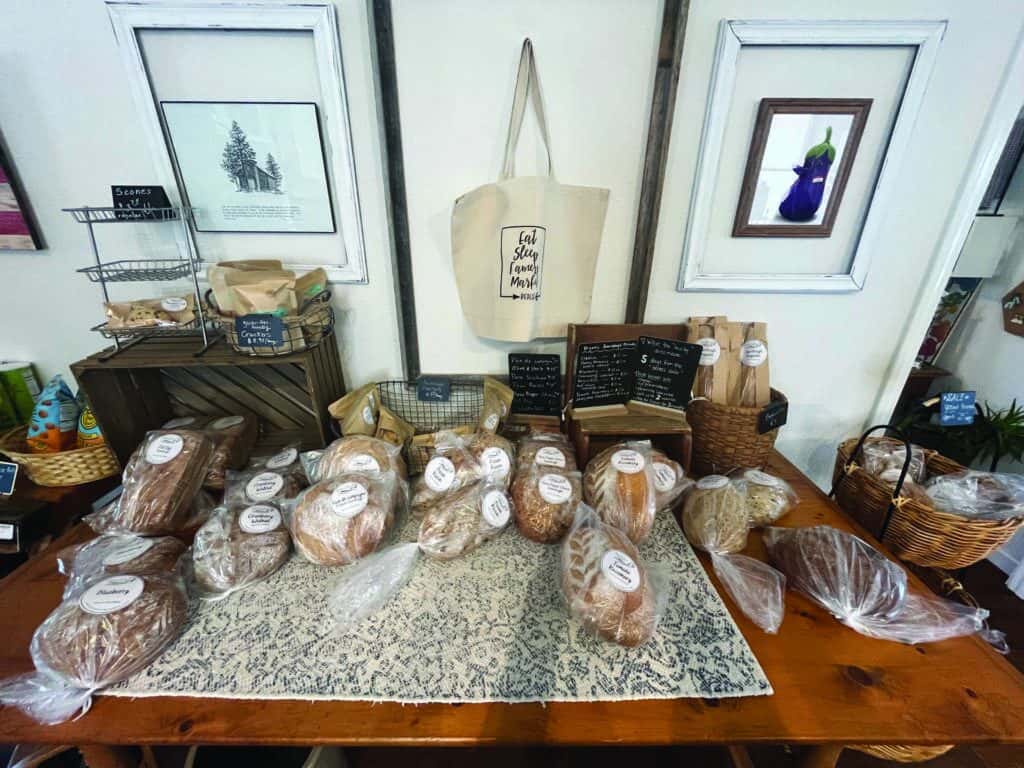 Various loaves of bread and baked goods are displayed on a wooden table, each wrapped in plastic and labeled, with tote bags and framed art on the wall behind.