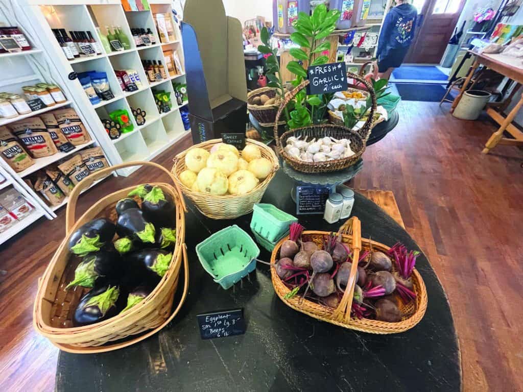 Vegetables and produce displayed in baskets on a wooden table inside a store, including eggplants, beets, onions, garlic, and leafy greens.
