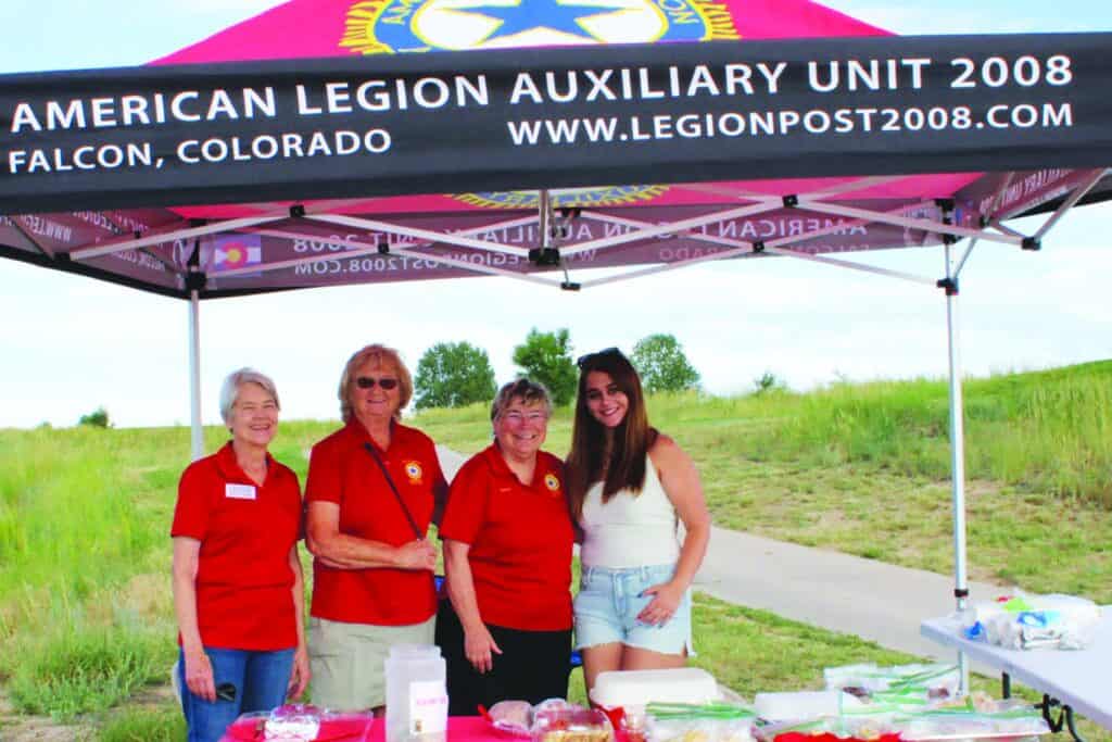 Four women stand under a canopy labeled "American Legion Auxiliary Unit 2008" with a table of food items in an outdoor grassy area.