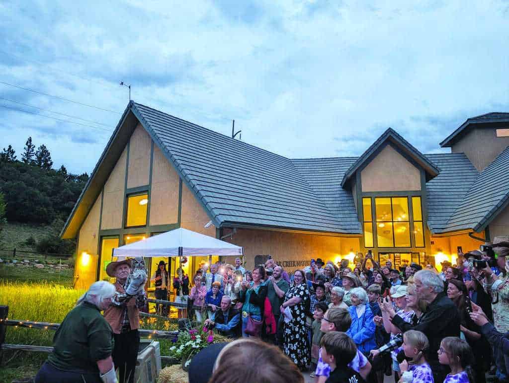 A large crowd gathers outside a house at dusk, watching a person holding an owl while others take photos and applaud.