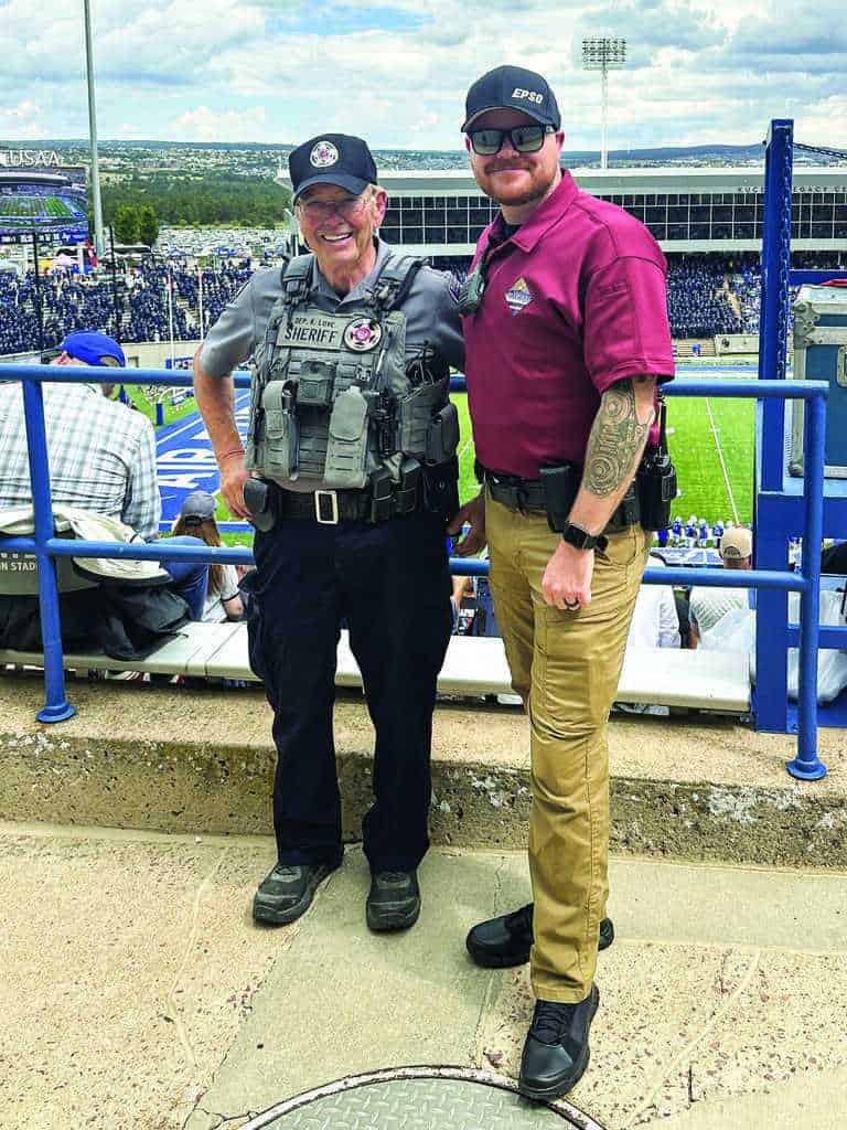 Two law enforcement officers, one in tactical gear and the other in a polo shirt, stand and smile at a sports stadium with a football field in the background.