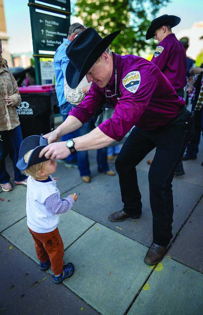 A man in a maroon uniform and cowboy hat places a black hat on a small child’s head during an outdoor event; other people stand in the background.