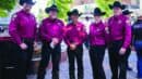Five people wearing matching maroon shirts, black pants, and black cowboy hats stand together outdoors, smiling at the camera.