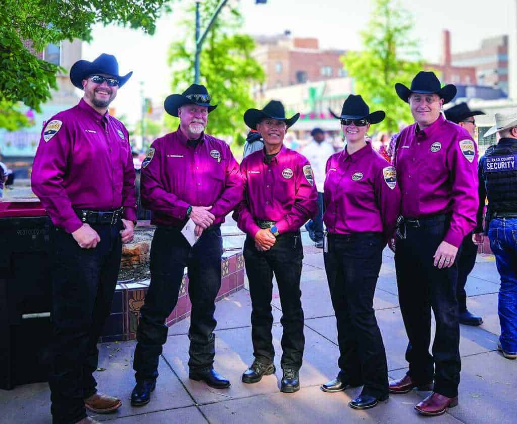 Five people wearing matching maroon shirts, black pants, and black cowboy hats stand together outdoors, smiling at the camera.