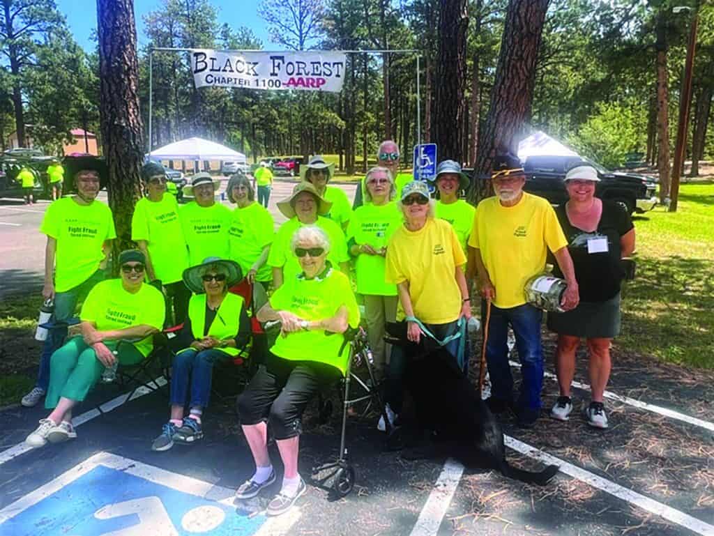 A group of people in yellow shirts pose together outdoors under a “Black Forest Chapter 1100 - AARP” banner at a park event.