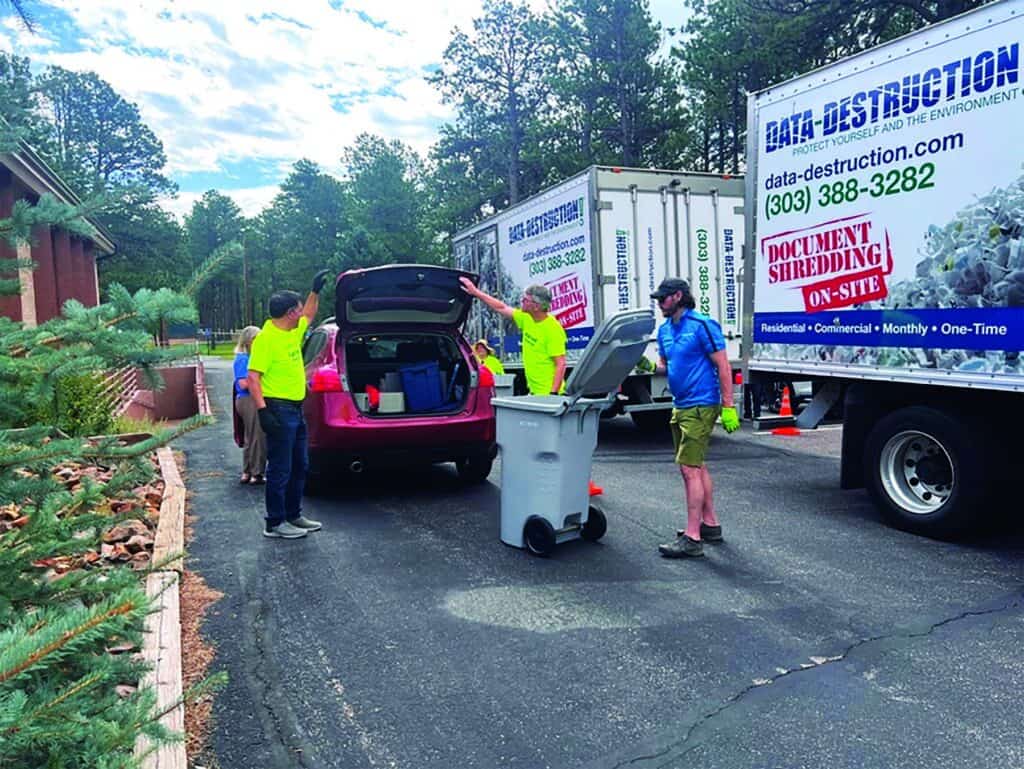People unload documents from a car into a bin for shredding near a document destruction truck in a parking lot surrounded by trees.