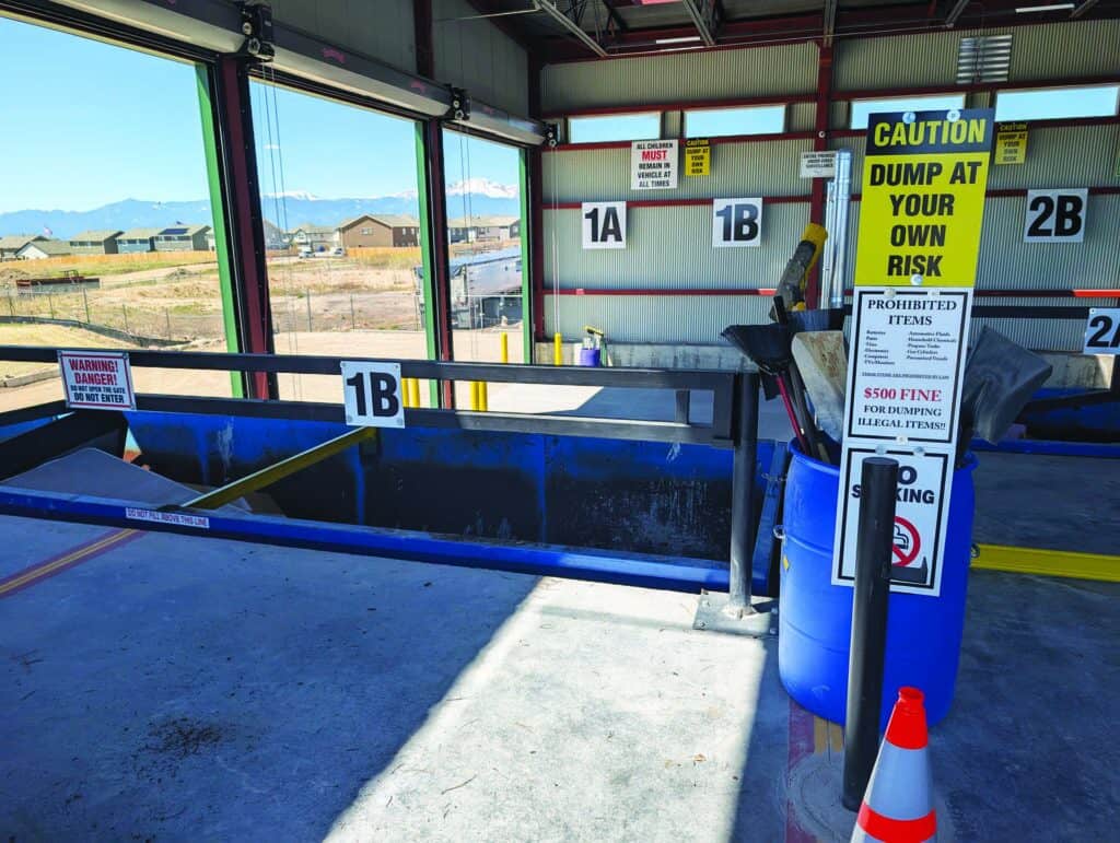 Indoor waste disposal area with labeled bins, caution signs, a blue barrel, and an orange cone; mountains and buildings visible through open wall.