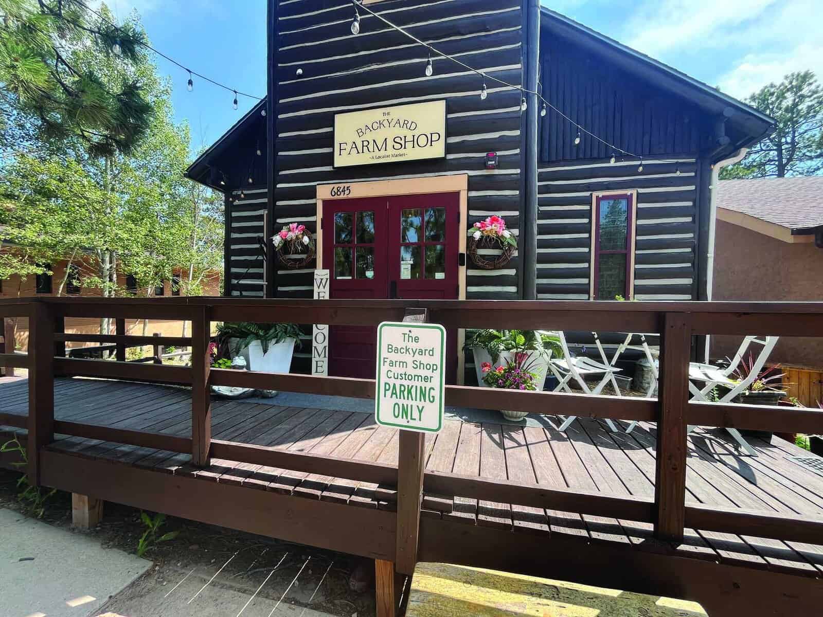 A wooden building with a sign reading "The Backyard Farm Shop" has a deck with potted flowers and a "Customer Parking Only" sign in front.