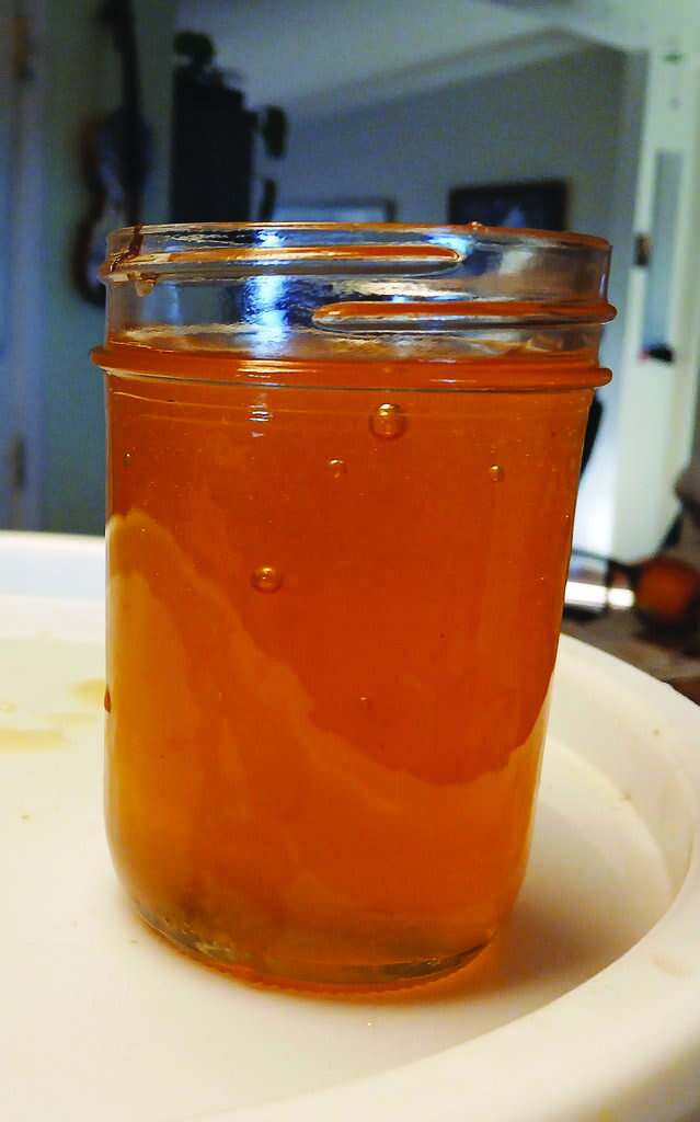 A glass jar filled with golden honey sits on a white surface indoors, with some honey residue visible along the inside of the jar.
