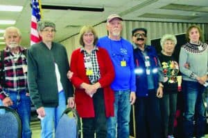 Seven older adults stand side by side indoors, some smiling, in a room with tables and an American flag in the background.