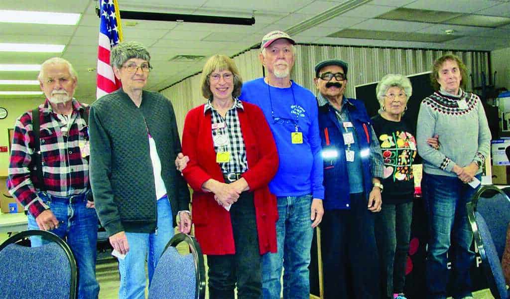 Seven older adults stand side by side indoors, some smiling, in a room with tables and an American flag in the background.
