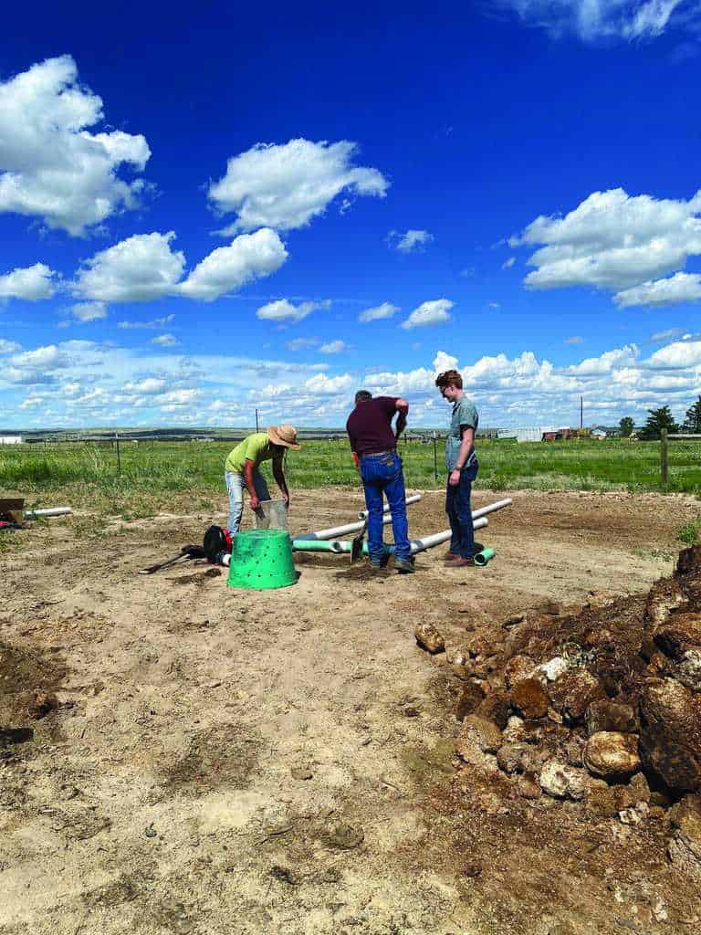 Three people stand on bare ground assembling or inspecting equipment under a blue sky with scattered clouds in a rural, grassy area.
