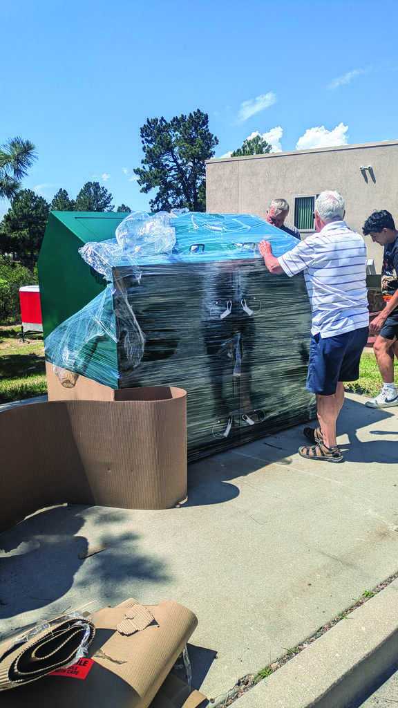 Three people unwrap and inspect a large stack of glass or acrylic panels on a pallet outside, near a building on a sunny day.