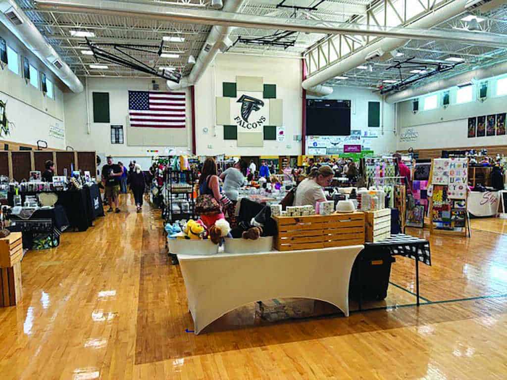 Vendors and shoppers are inside a school gymnasium, with tables displaying various goods. An American flag and a "Falcons" sign are visible on the wall.