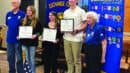 Five people stand indoors; three young individuals hold certificates while flanked by two older adults. Exchange Club banners and an American flag are visible in the background.