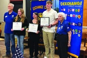 Five people stand indoors; three young individuals hold certificates while flanked by two older adults. Exchange Club banners and an American flag are visible in the background.
