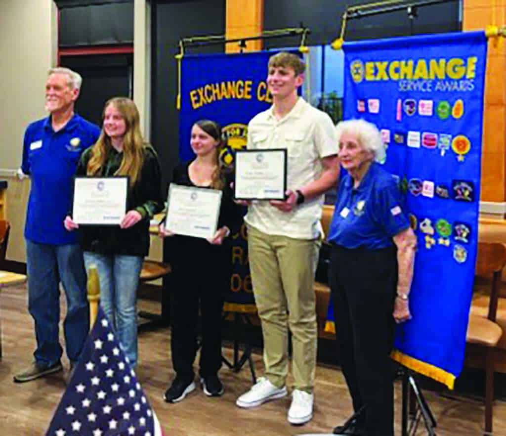 Five people stand indoors; three young individuals hold certificates while flanked by two older adults. Exchange Club banners and an American flag are visible in the background.