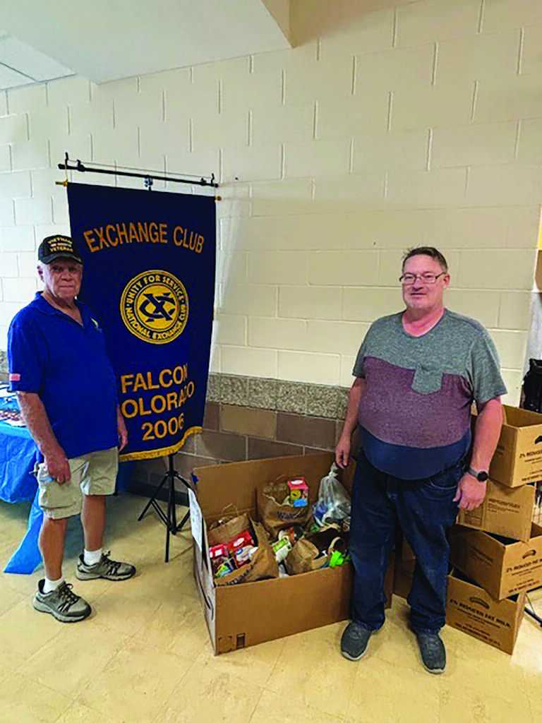 Two men stand beside boxes of donated food items in front of an Exchange Club of Falcon, Colorado banner inside a building.