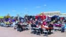 People sit at tables and gather around food trucks at an outdoor food festival on a sunny day with clear skies.