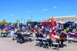 People sit at tables and gather around food trucks at an outdoor food festival on a sunny day with clear skies.