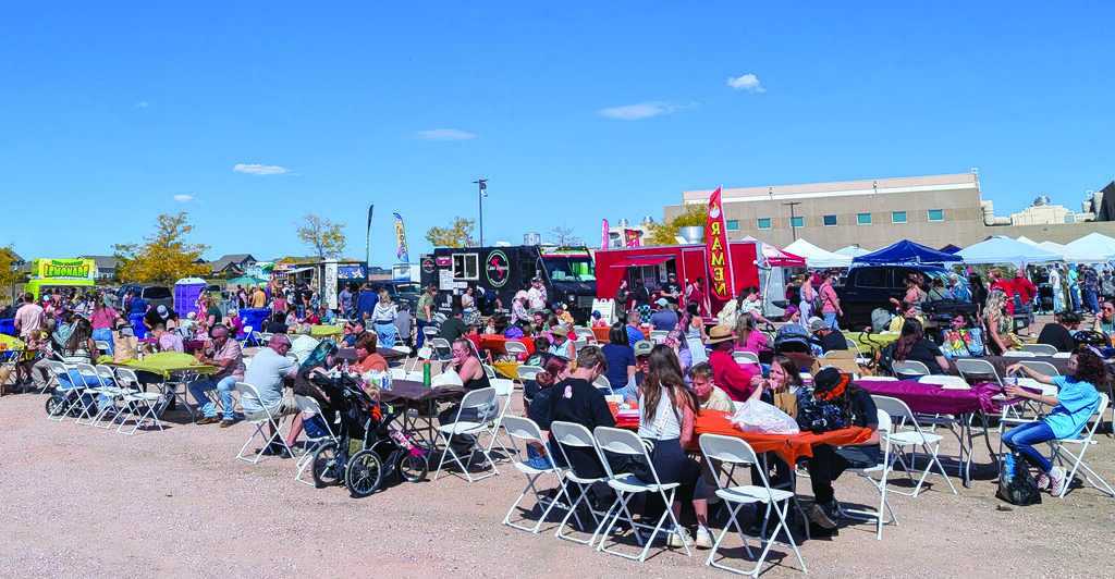 People sit at tables and gather around food trucks at an outdoor food festival on a sunny day with clear skies.