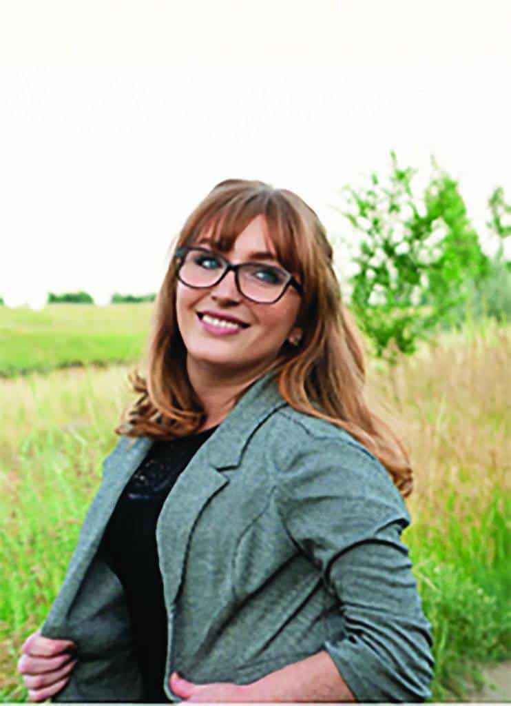 A woman with long light brown hair and glasses wearing a gray blazer stands outdoors in a grassy field, smiling at the camera.