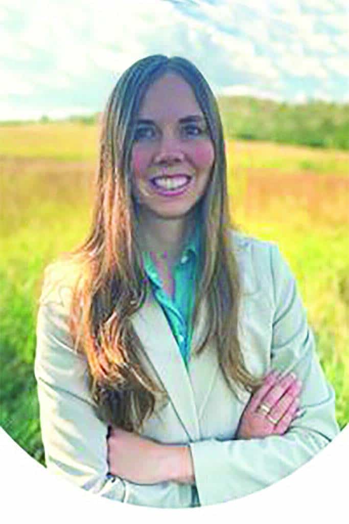 A woman with long brown hair, wearing a light-colored blazer and green shirt, stands outdoors in a grassy field with her arms crossed, smiling at the camera.