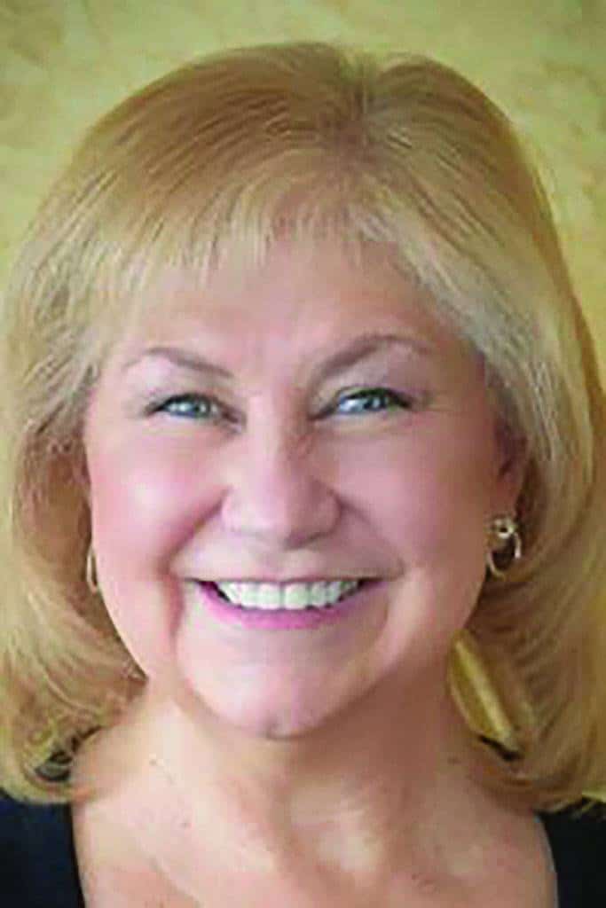 A middle-aged woman with blonde hair smiles at the camera, wearing hoop earrings and a dark top, against a light yellow background.