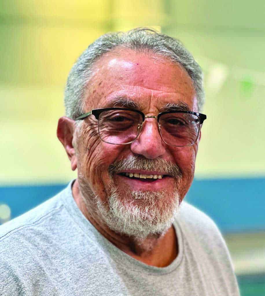 An older man with gray hair, a beard, and glasses is smiling while wearing a light gray shirt. The background is softly blurred.