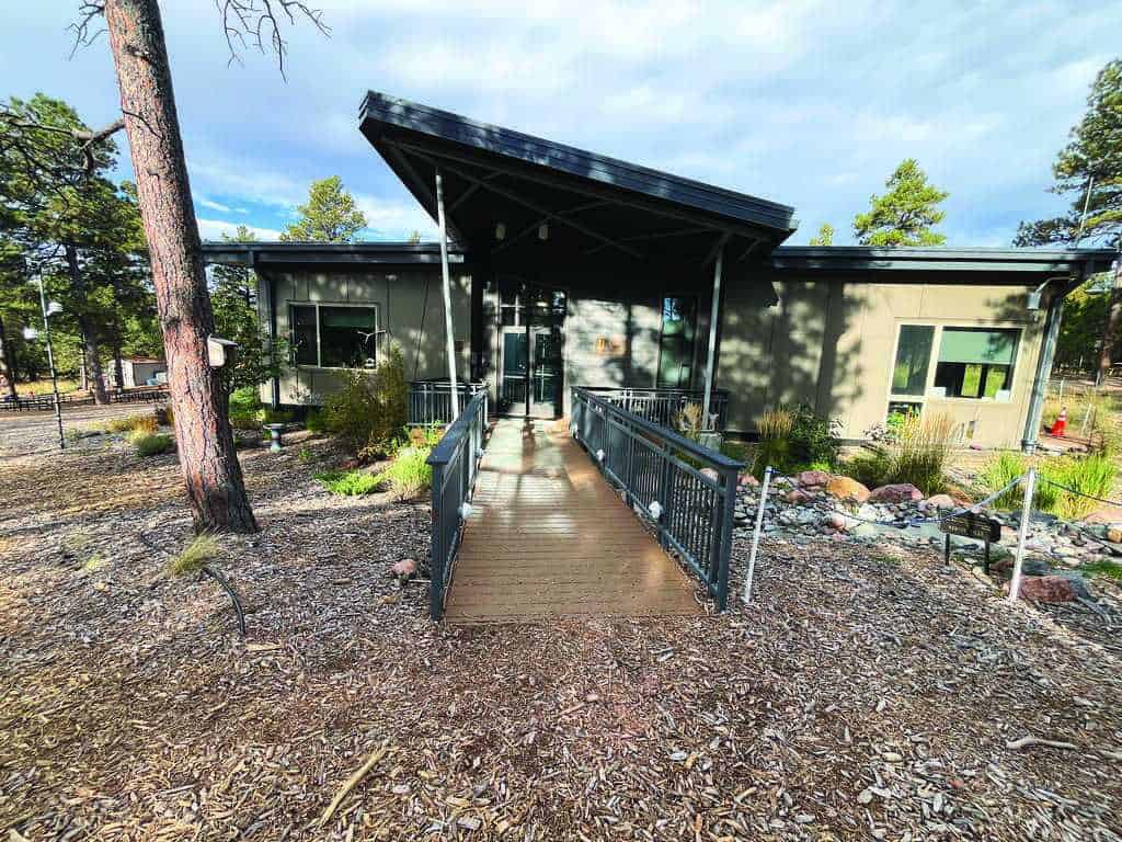 A modern, single-story building with large windows, a slanted roof, and a ramp entrance, surrounded by trees and wood chip landscaping.