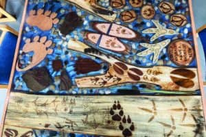 A wooden table decorated with animal tracks, paw prints, and natural elements set in resin with blue and stone accents.