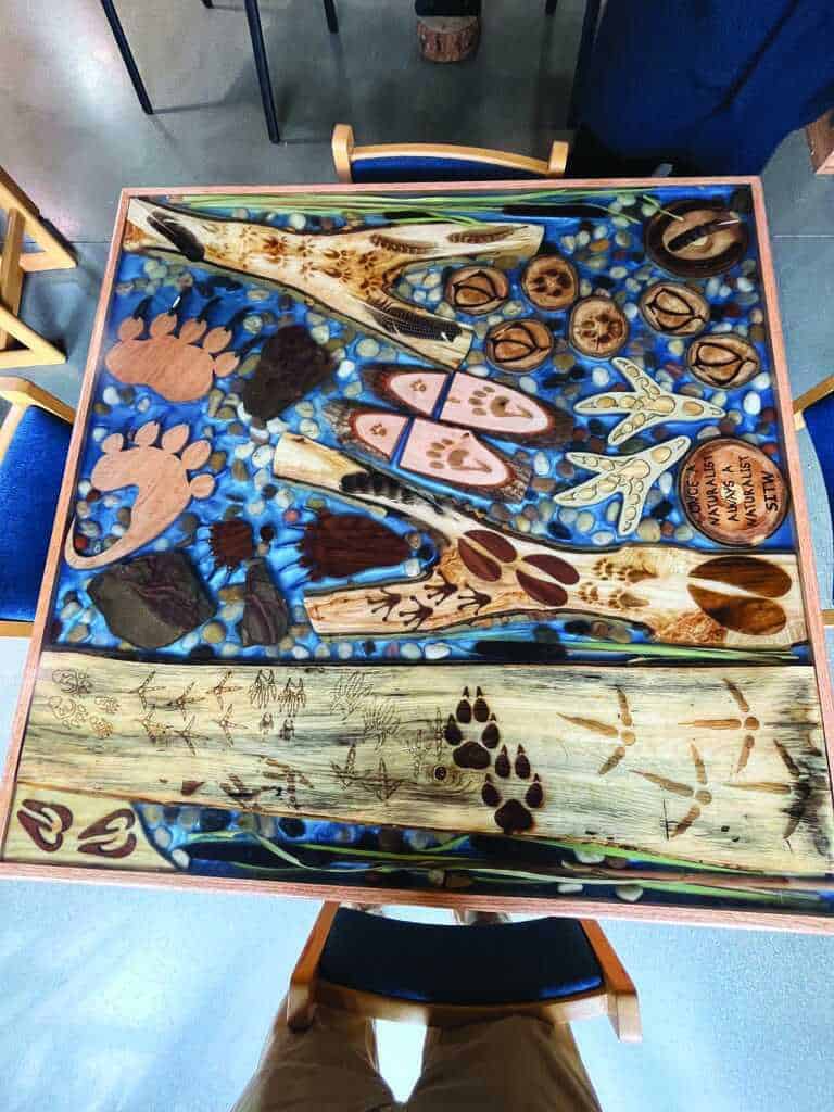 A wooden table decorated with animal tracks, paw prints, and natural elements set in resin with blue and stone accents.