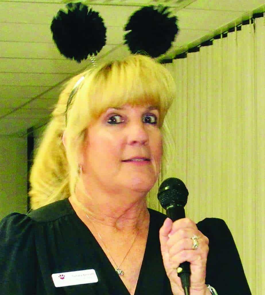 A woman with blonde hair wearing a headband with two black pom-poms speaks into a microphone indoors. She is wearing a black top and a name badge.