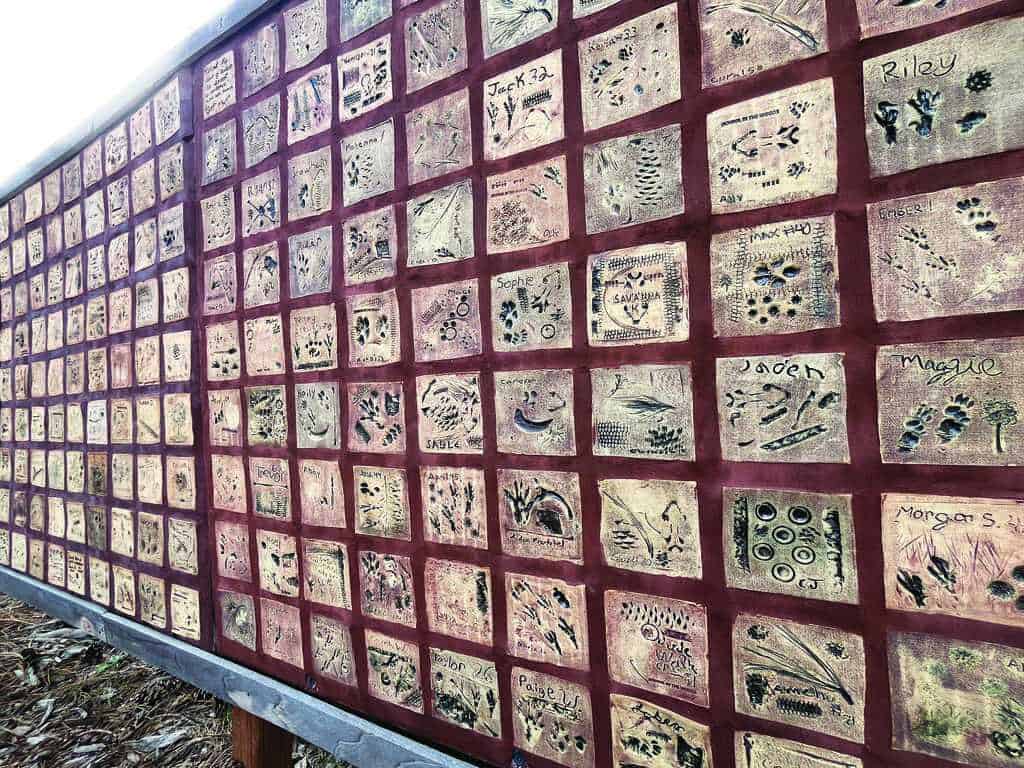 A wall display of individual ceramic tiles, each featuring unique imprints, drawings, or names, arranged in a grid pattern with a red border between tiles.