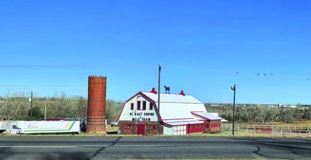 A red and white barn with a silo stands next to a road under a clear blue sky. The barn has "AL KALY SHRINE" written on the front.