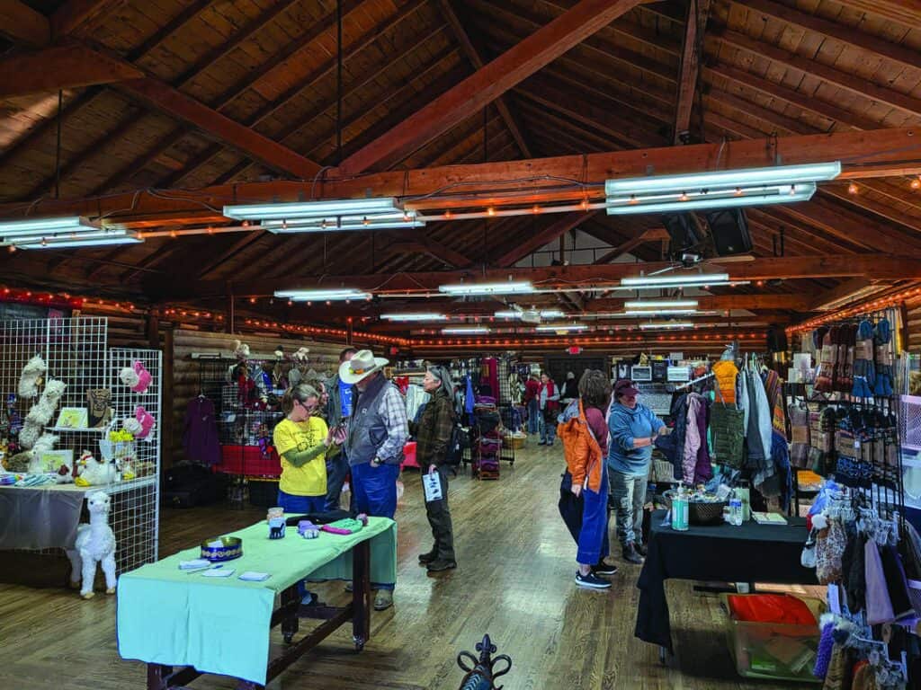 People browse various vendor booths inside a large, wooden hall with exposed beams and string lights; tables display crafts, clothing, and goods.