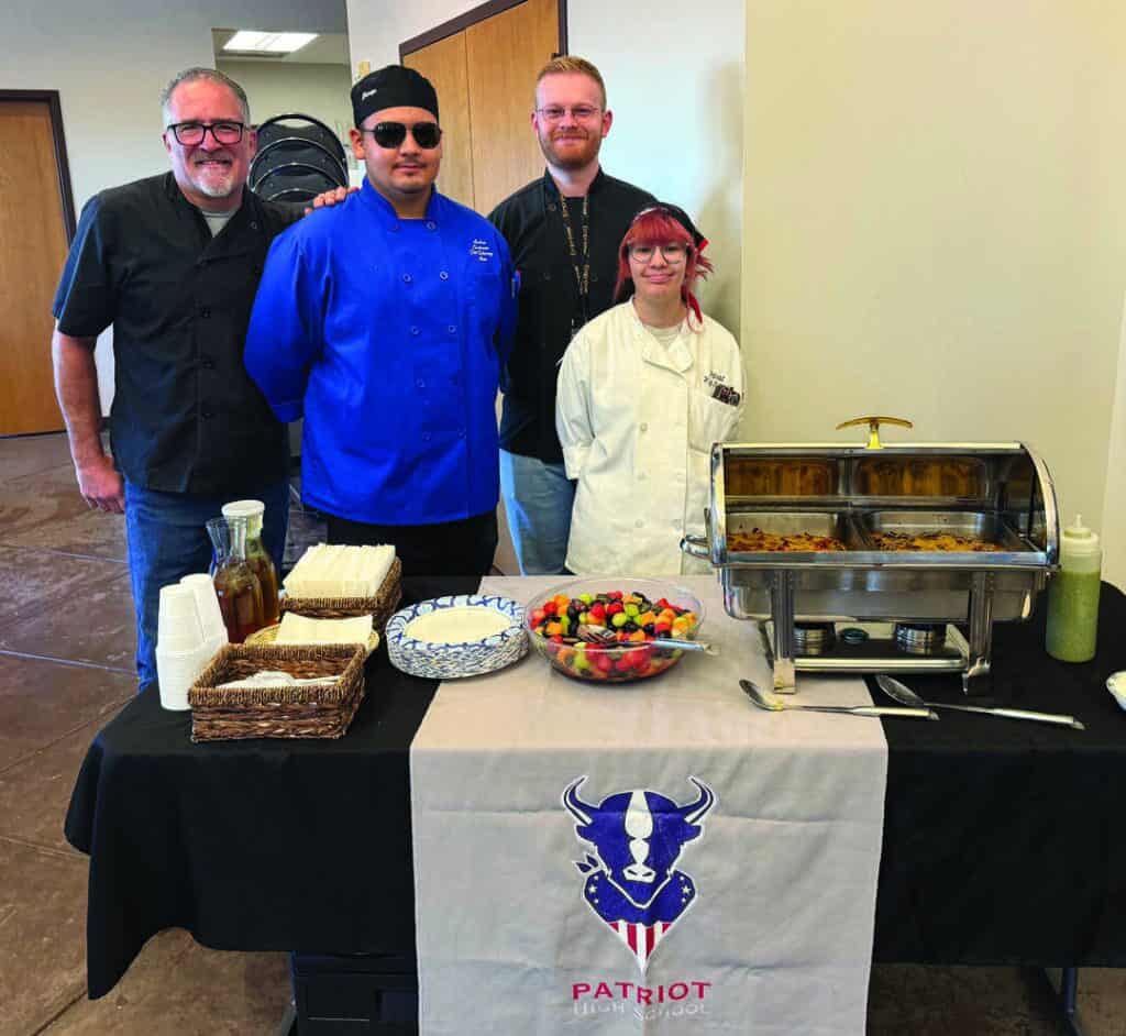 Four people stand behind a catering table with food, fruit, and condiments; a "Patriot High School" banner hangs on the table.