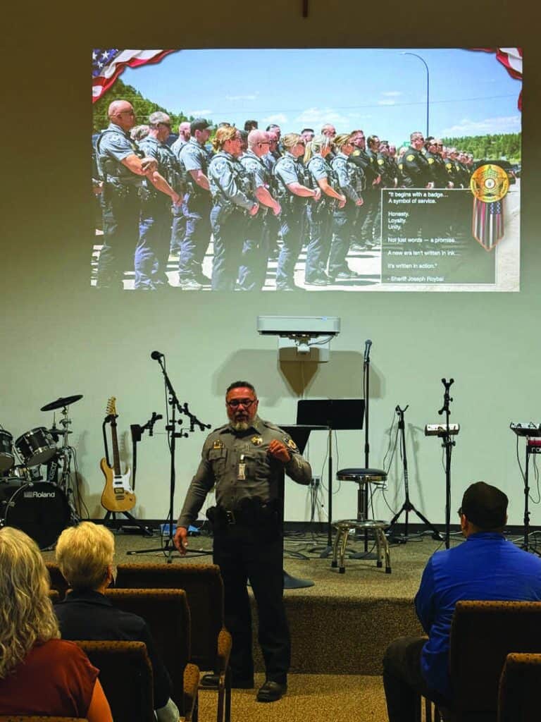 A police officer speaks to an audience in a church while a projected image of uniformed officers standing in a row is displayed behind him.