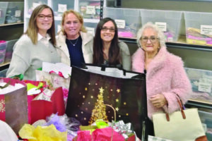 Four women stand in front of shelves with bins, surrounded by colorful gift bags and tissue paper in a storage room.