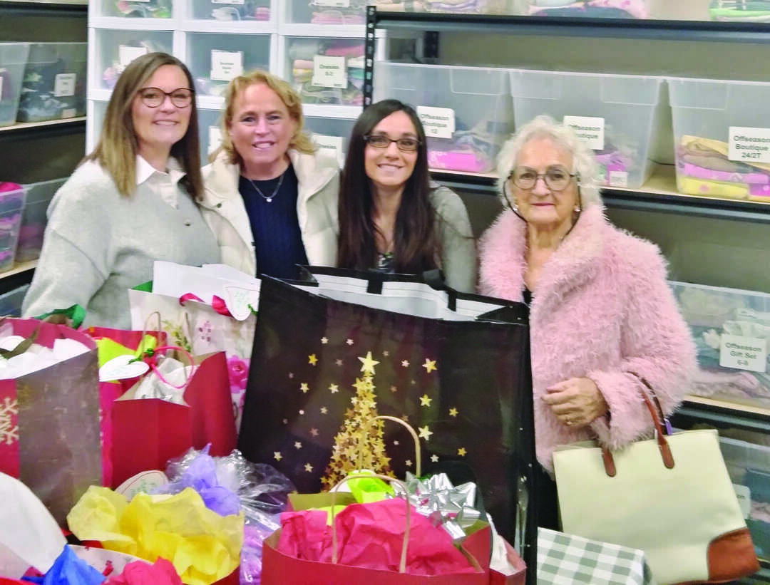 Four women stand in front of shelves with bins, surrounded by colorful gift bags and tissue paper in a storage room.