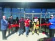 Six people, including one in a firefighter mascot costume, stand in front of a building cutting a red ribbon for a ceremonial grand opening event.