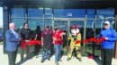 Six people, including one in a firefighter mascot costume, stand in front of a building cutting a red ribbon for a ceremonial grand opening event.