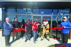 Six people, including one in a firefighter mascot costume, stand in front of a building cutting a red ribbon for a ceremonial grand opening event.