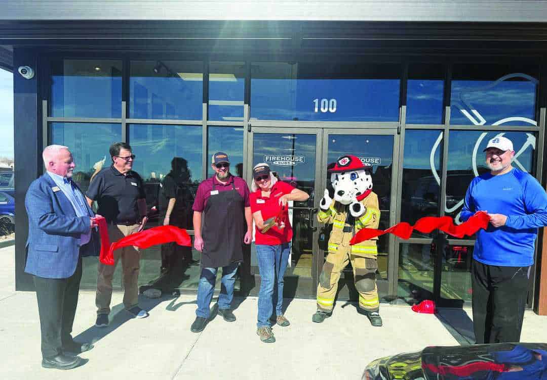Six people, including one in a firefighter mascot costume, stand in front of a building cutting a red ribbon for a ceremonial grand opening event.