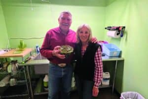 Two adults stand together in a green room near a sink and counter; one is holding a bowl of salad greens, both are smiling at the camera.