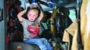 A young boy sits inside a fire truck, wearing a t-shirt with a car graphic and holding a firefighter helmet on his head.