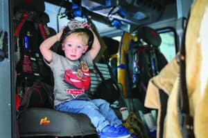 A young boy sits inside a fire truck, wearing a t-shirt with a car graphic and holding a firefighter helmet on his head.