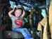 A young boy sits inside a fire truck, wearing a t-shirt with a car graphic and holding a firefighter helmet on his head.