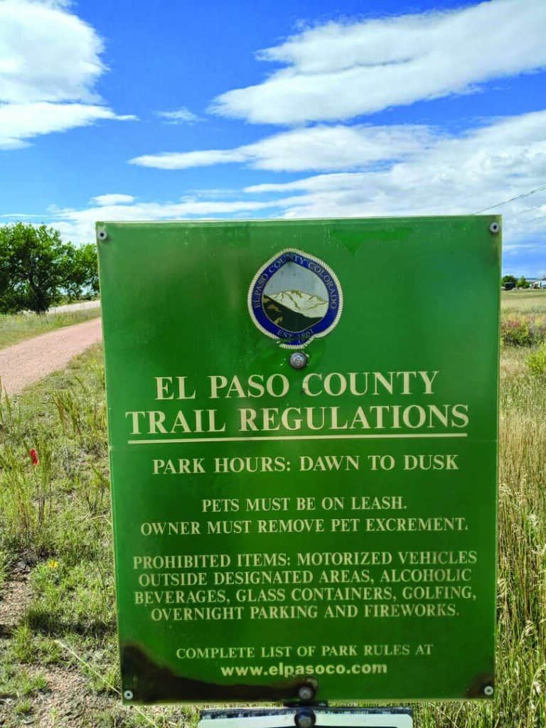 Green sign listing El Paso County trail regulations, including park hours, pet rules, and prohibited items like vehicles, alcohol, glass, golfing, overnight parking, and fireworks.