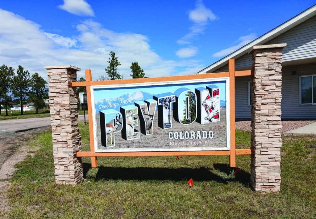 A large welcome sign reads "Greetings from Peyton, Colorado, Elevation 6816 ft," mounted between stone pillars, with a building and trees in the background.
